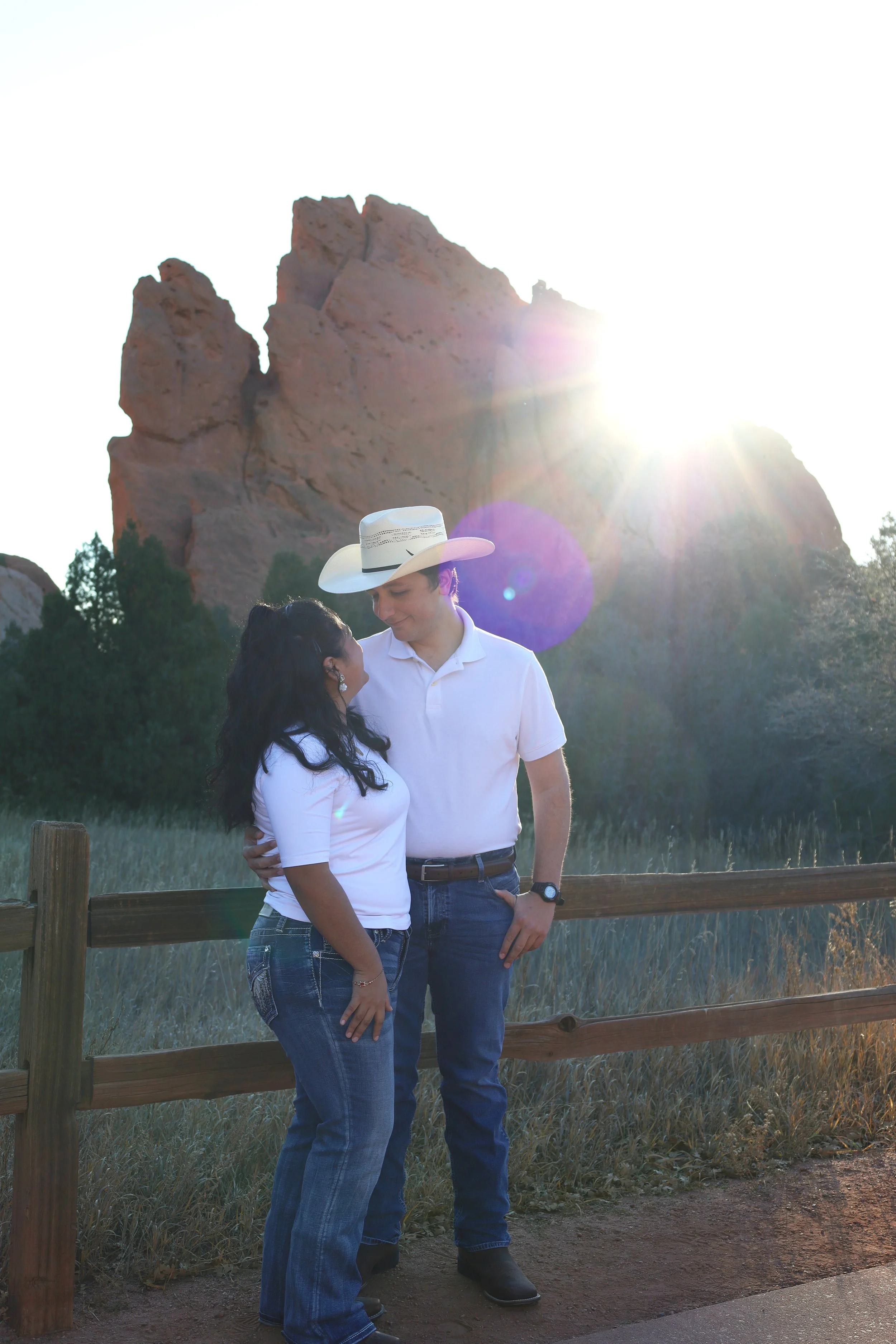 A couple stands close together, smiling and looking into each other's eyes, with a large rock formation and the sun shining brightly behind them in the background.