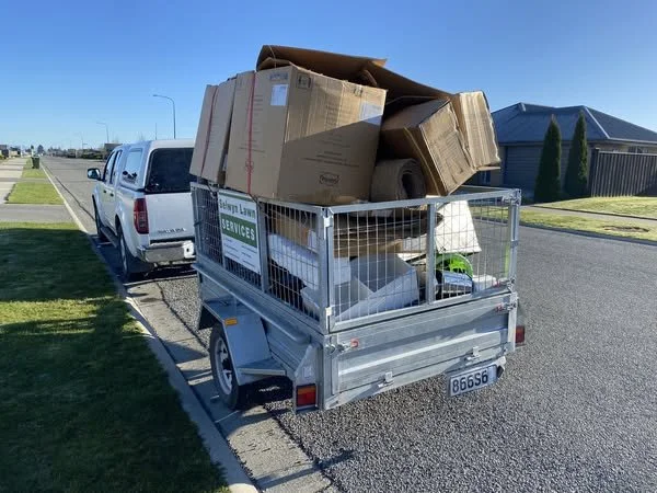 A small utility trailer filled with large cardboard boxes and various packing materials, parked on the side of a suburban street with a white pickup truck attached. There are houses and a clear blue sky in the background.