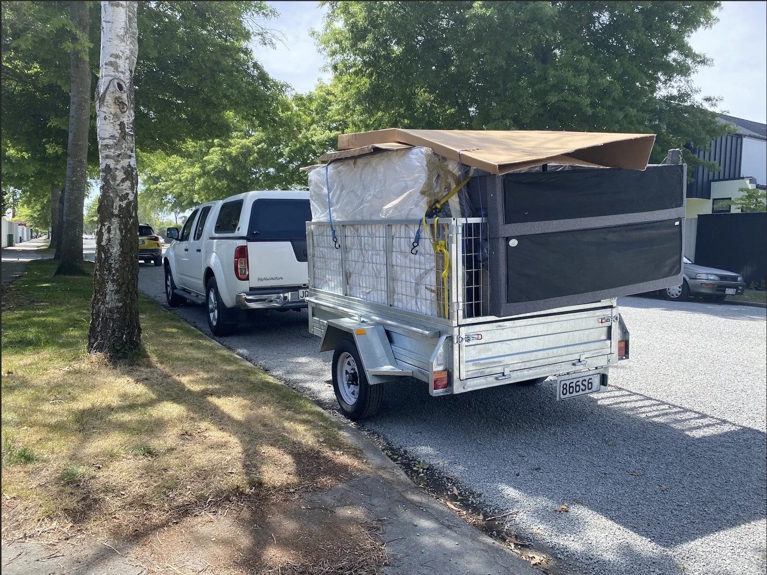 A small trailer parked on the side of a tree-lined street, loaded with various bags and boxes secured with straps and cover.