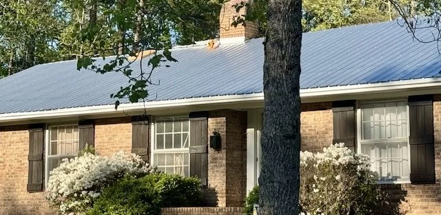 A brick house with black shutters, white window frames, and a metal roof, surrounded by trees and bushes.