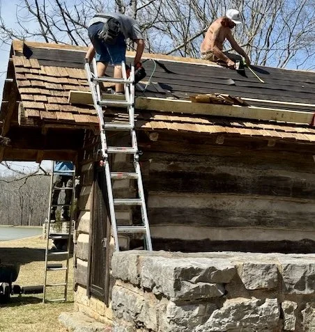 Two workers on a roof repairing shingles, using ladders for access, during daytime with leafless trees in the background.