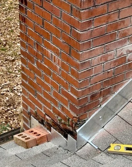 Close-up of a brick chimney corner outside a building, with metal flashing installed at the base and several bricks on the ground nearby.