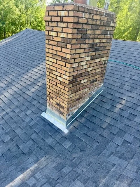 Close-up of a brick chimney on a shingled roof with a metal flashing installed at the base.