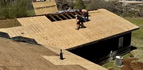 Two workers on a house roof under construction, with plywood sheathing in place and a hole cut out for a future skylight or window.
