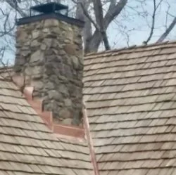 A stone chimney extending above a shingled roof with a chimney cap and a chimney brush inside