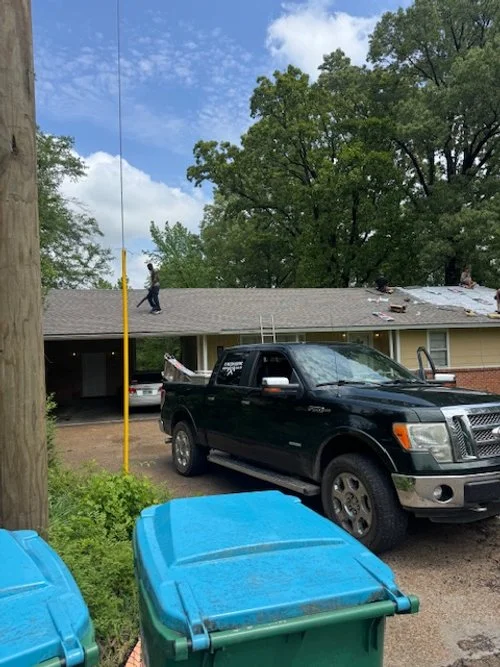 A man on the roof of a single-story house working near the roof's edge with roofing tools. A black pickup truck is parked in the driveway, and two blue trash bins are in the foreground. Tall trees and a partly cloudy sky are in the background.