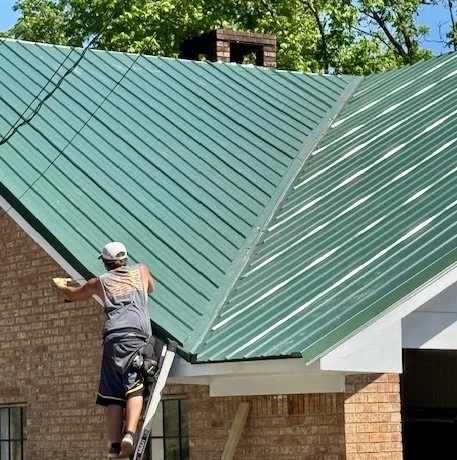 A person standing on a ladder working on installing a green metal roof on a brick house, with trees in the background.