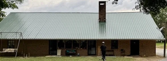 A person standing in front of a brick house with a green metal roof, trees on the sides, and a front porch.