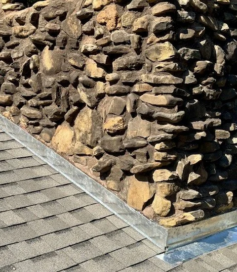 Close-up of a stone chimney on a house with a roof in the foreground.