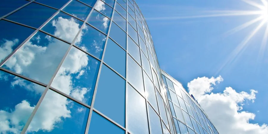 A modern glass office building reflecting clouds and blue sky, with sunlight shining overhead.