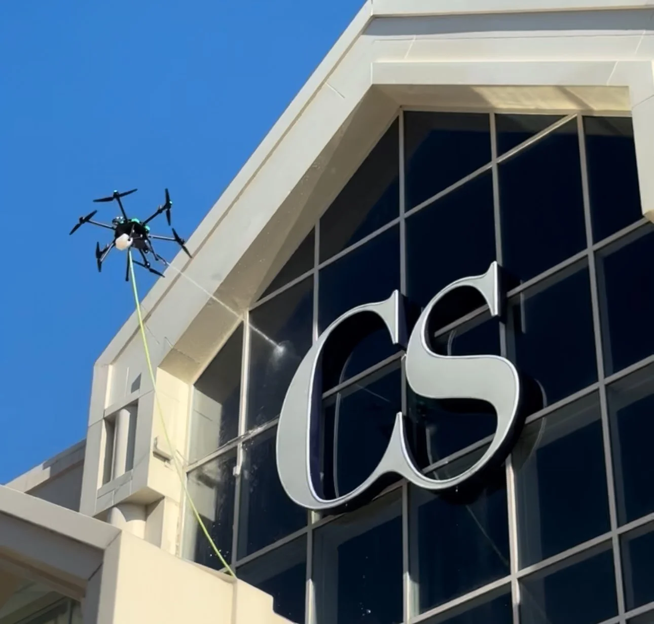 A drone is spraying disinfectant on the exterior of a building with a large sign displaying the letters 'CS' in front of a glass window.