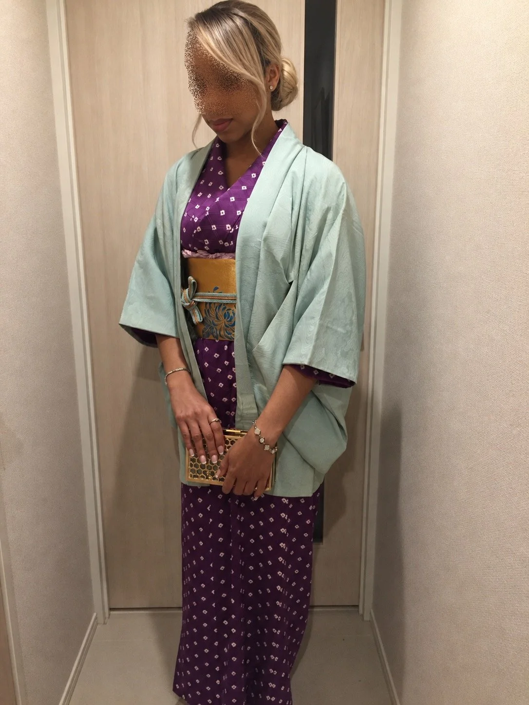 Woman wearing a purple patterned kimono-style dress, mint green haori jacket, decorative obi belt, and holding a gold clutch