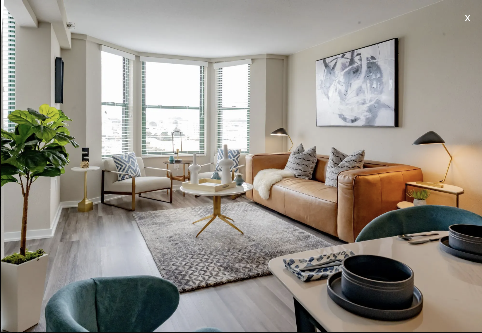 Living room with a large window, tan leather sofa, blue armchair, patterned pillows, abstract artwork, and a white round coffee table with vases and decorative items.