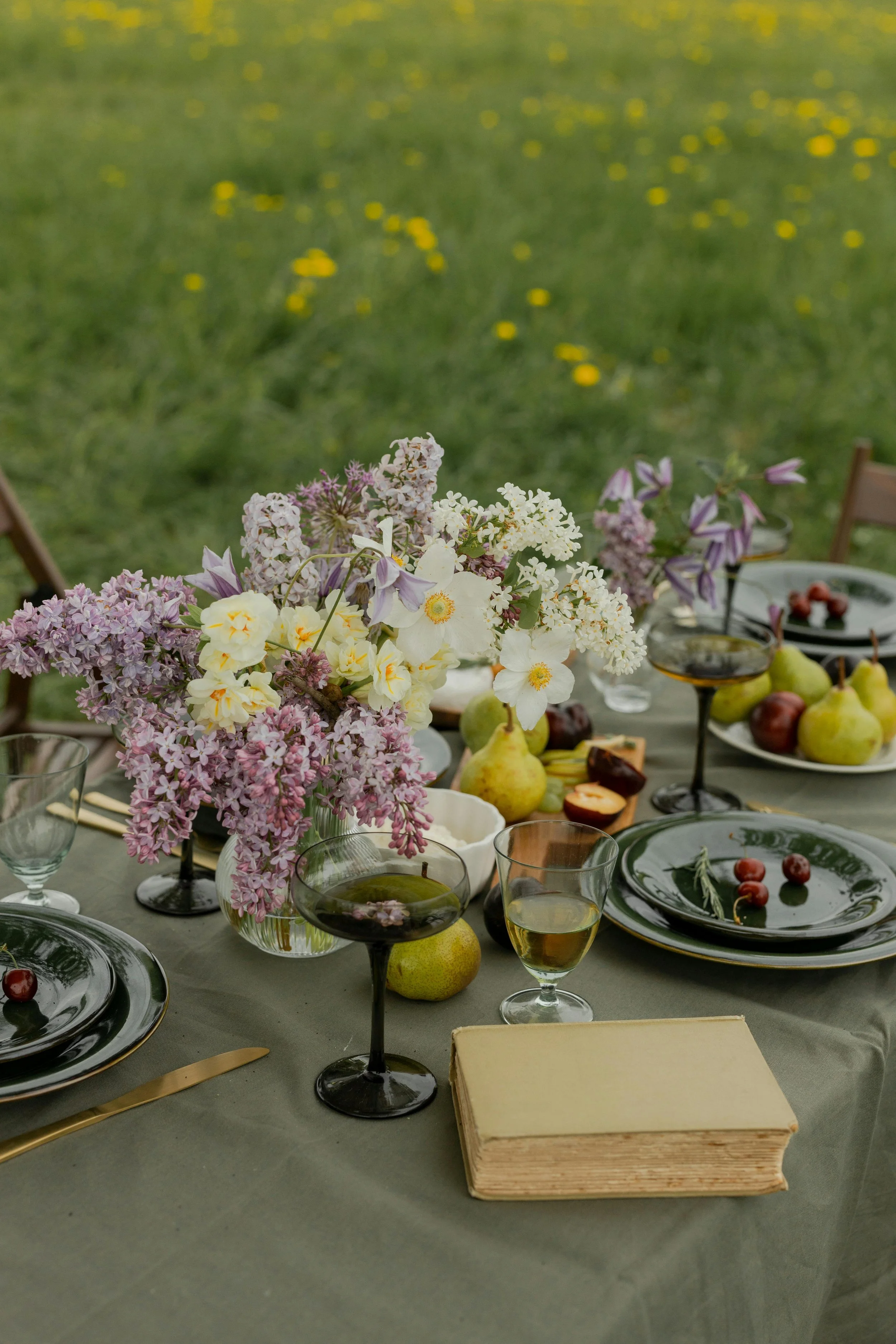 A table set outdoors with black plates, gold utensils, and glasses filled with wine and water. The table features a large floral centerpiece with purple, white, and yellow flowers, along with various fruits like pears, apples, and cherries, and a vintage book.