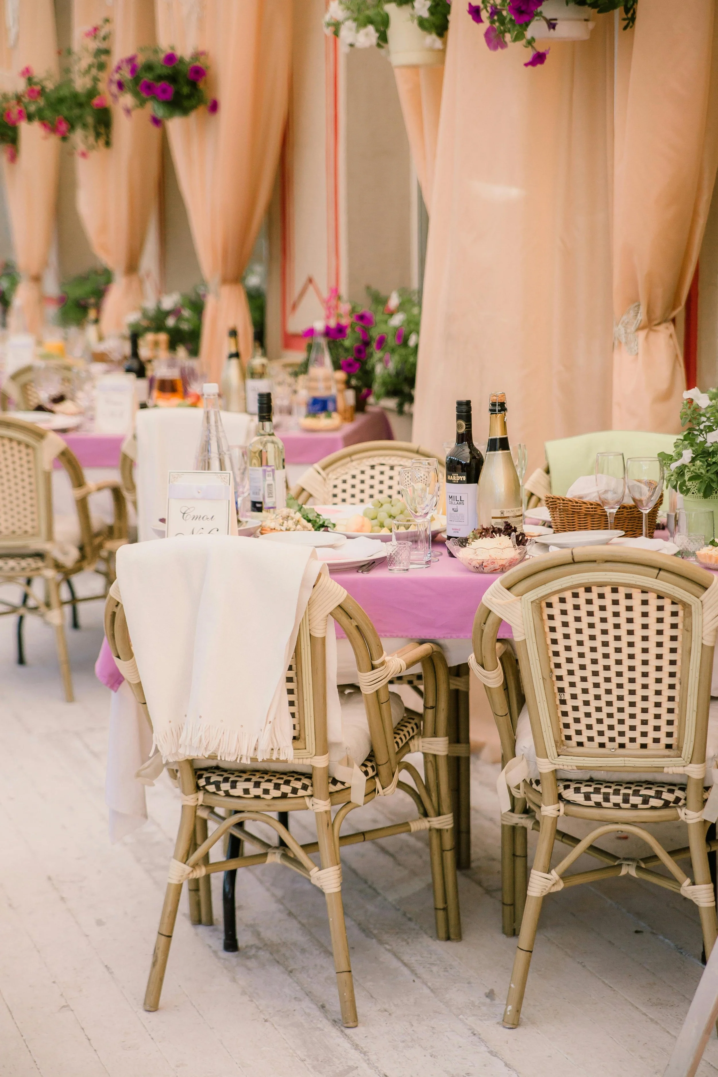 A decorated banquet table with pink tablecloths, wine bottles, glasses, plates, and floral centerpiece, set against pink curtains with hanging flower arrangements inside a bright room.