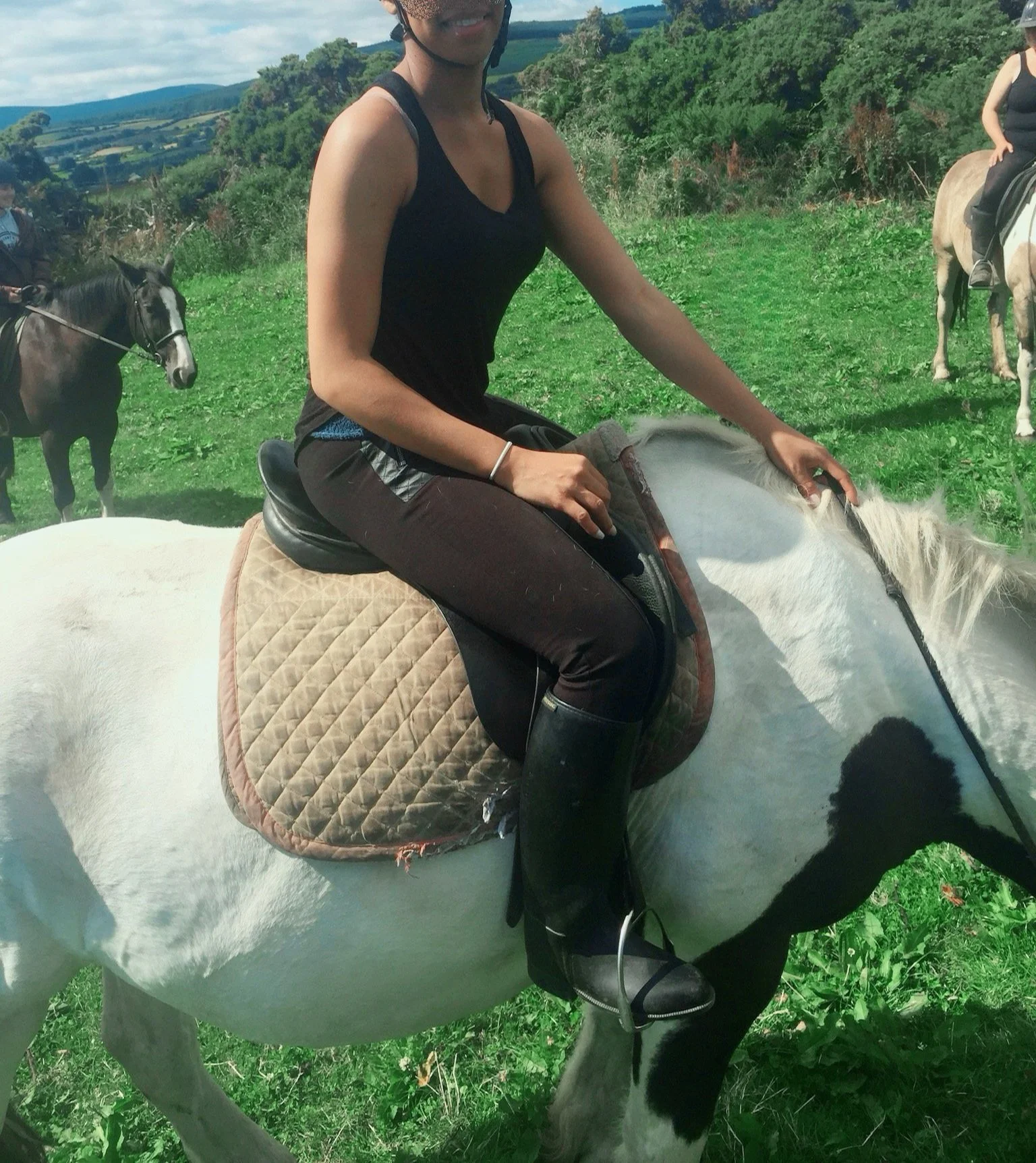 A woman riding a white horse with black patches, wearing a black tank top, brown riding pants, and black riding boots, with green pasture and other riders on horses in the background.