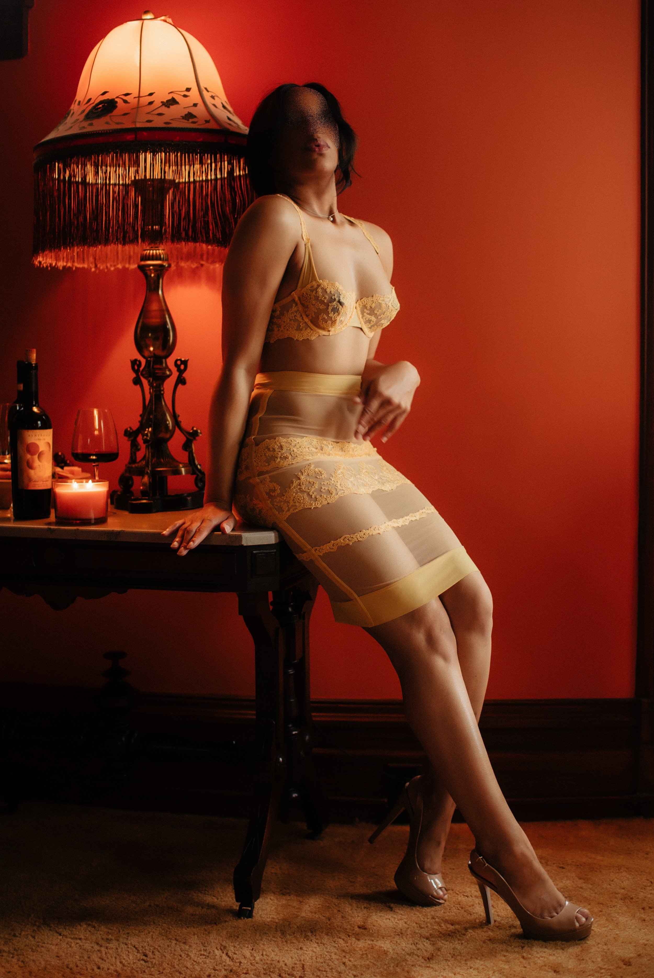A woman in yellow lace lingerie sitting on a table in a warmly lit room, with a red wall, a vintage lamp, and candles.