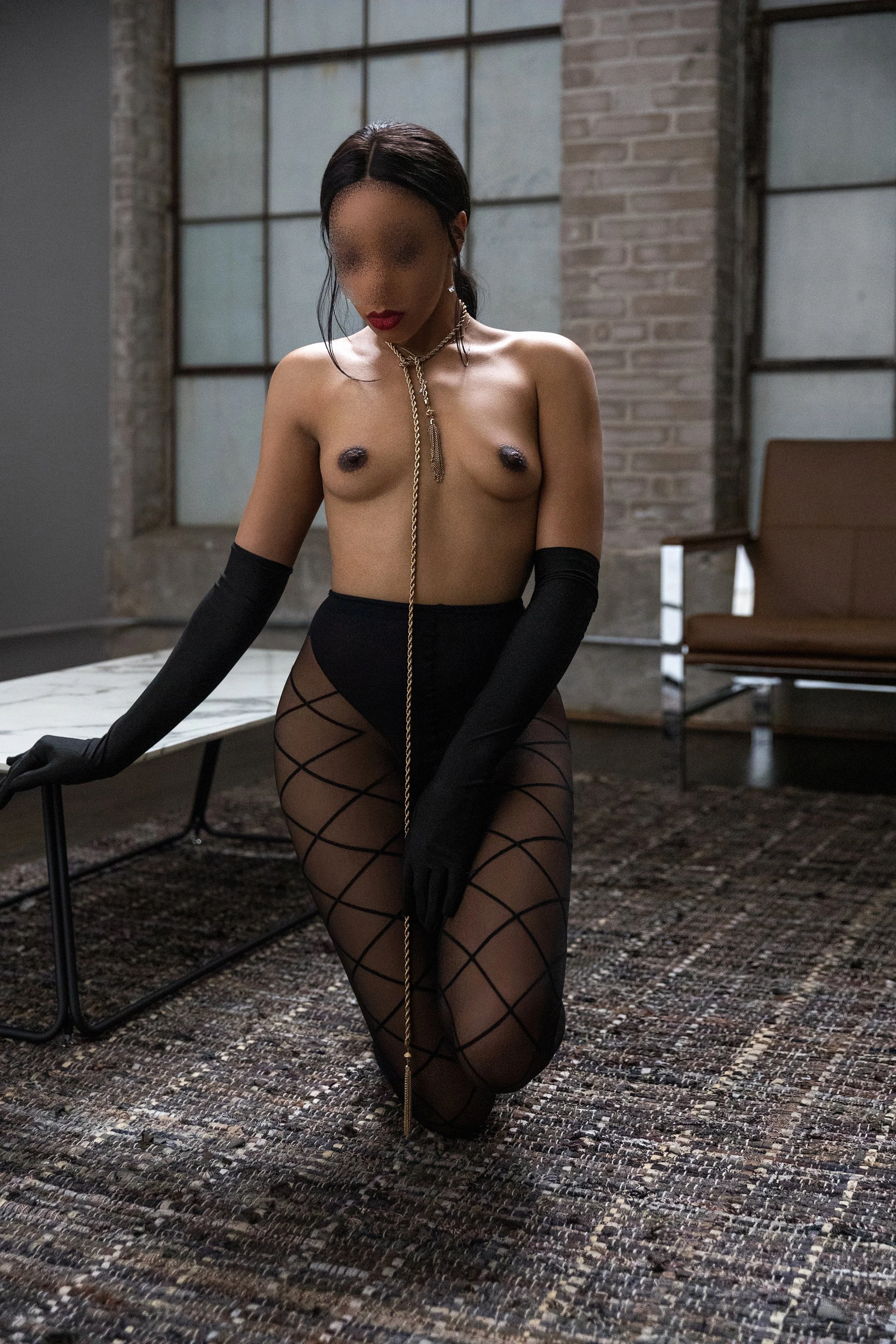 A topless woman wearing black fishnet stockings, long black gloves, and jewelry, kneeling on a patterned rug in an industrial-style room with large windows and brick walls.