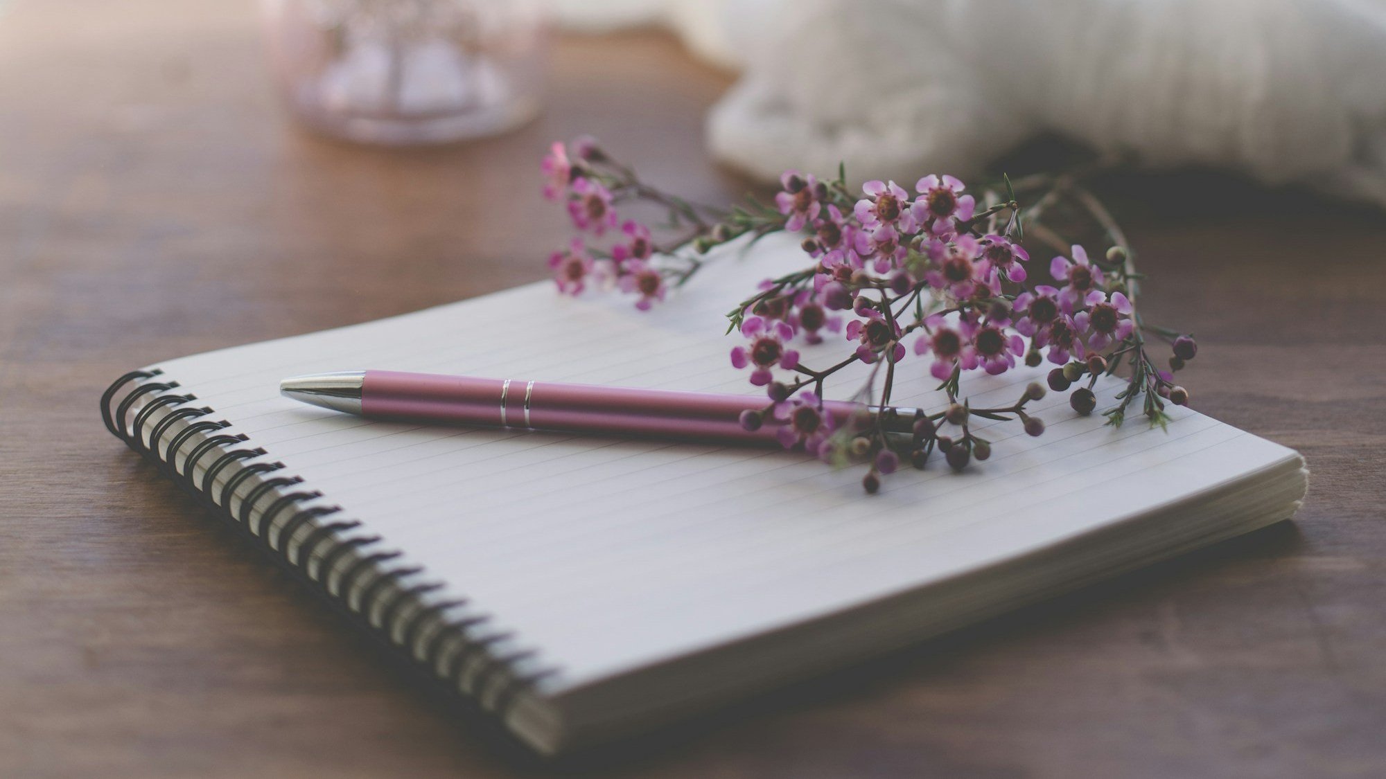 Open spiral notebook with lined pages, pink pen resting on it, and pink flowers on top, on a wooden surface.
