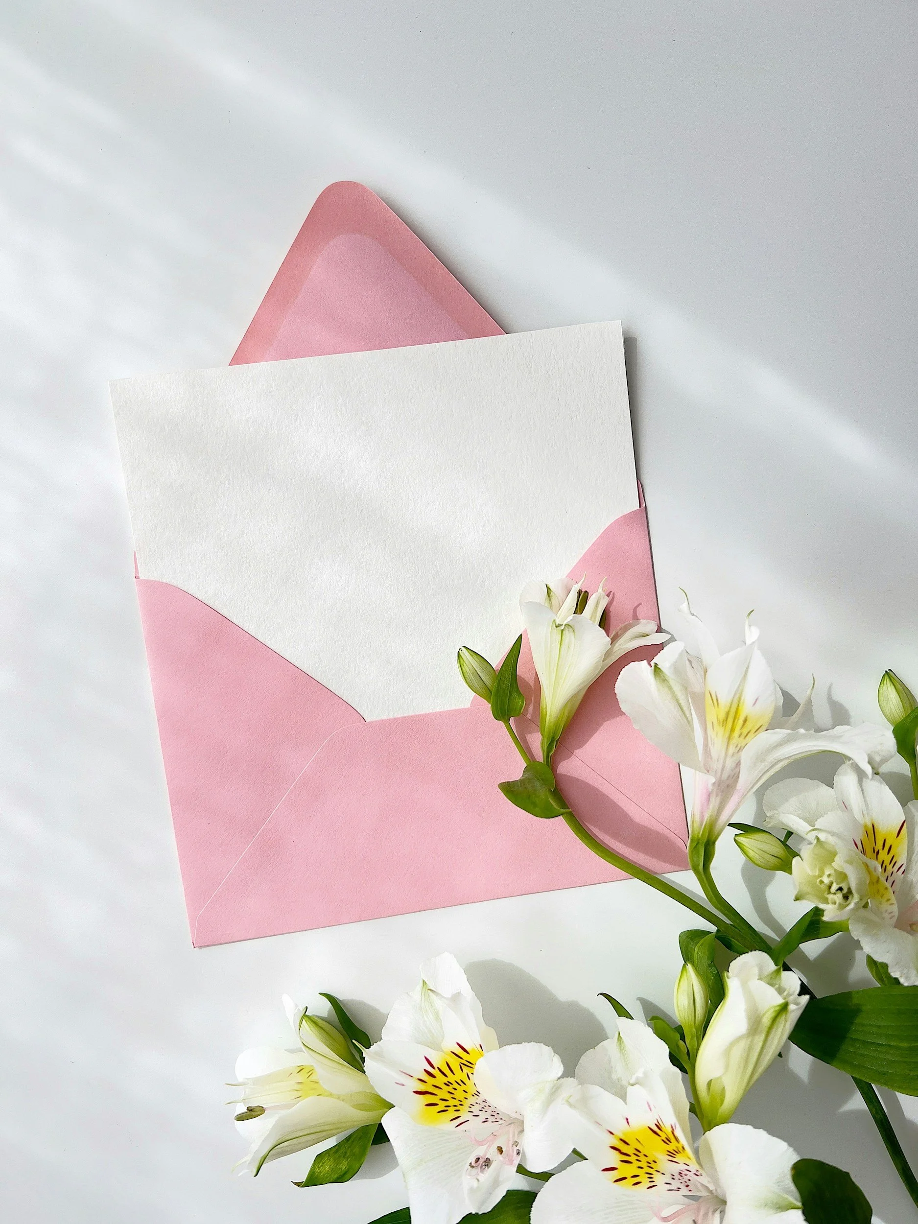 Open pink envelope with blank white note inside, surrounded by white lilies with yellow and maroon accents on a white surface.