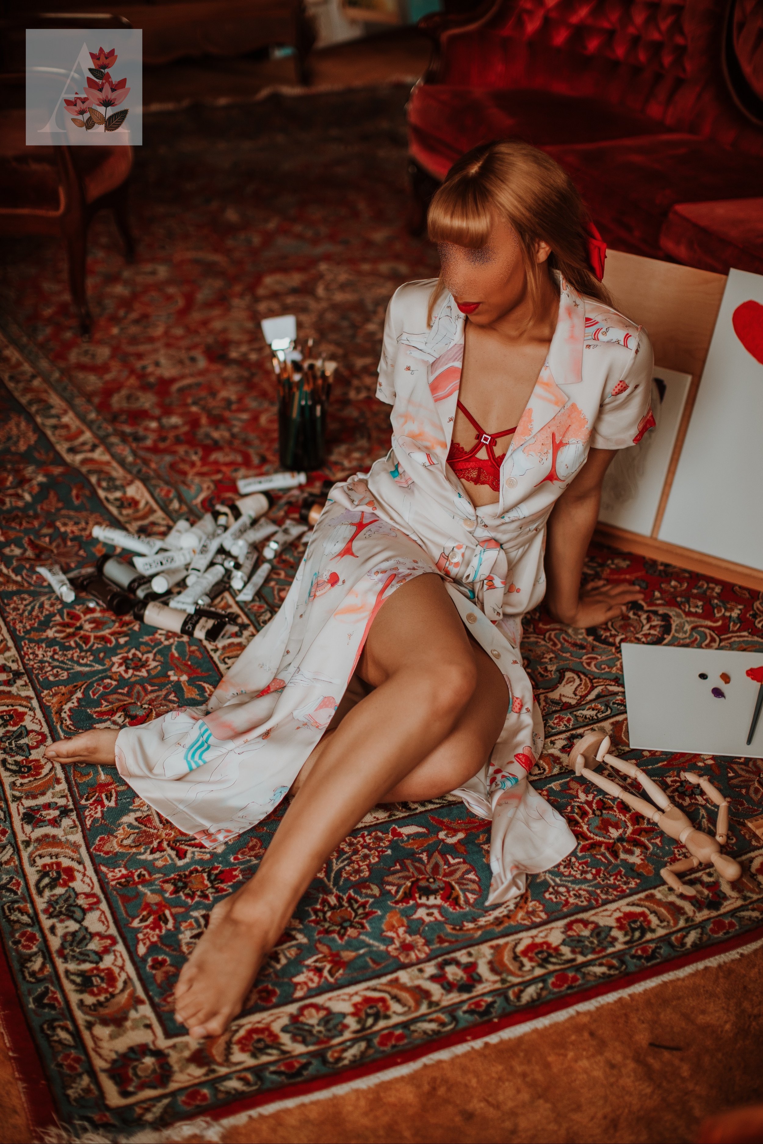 Woman in a patterned outfit sitting on a colorful rug beside art supplies, a canvas, and an artist mannequin