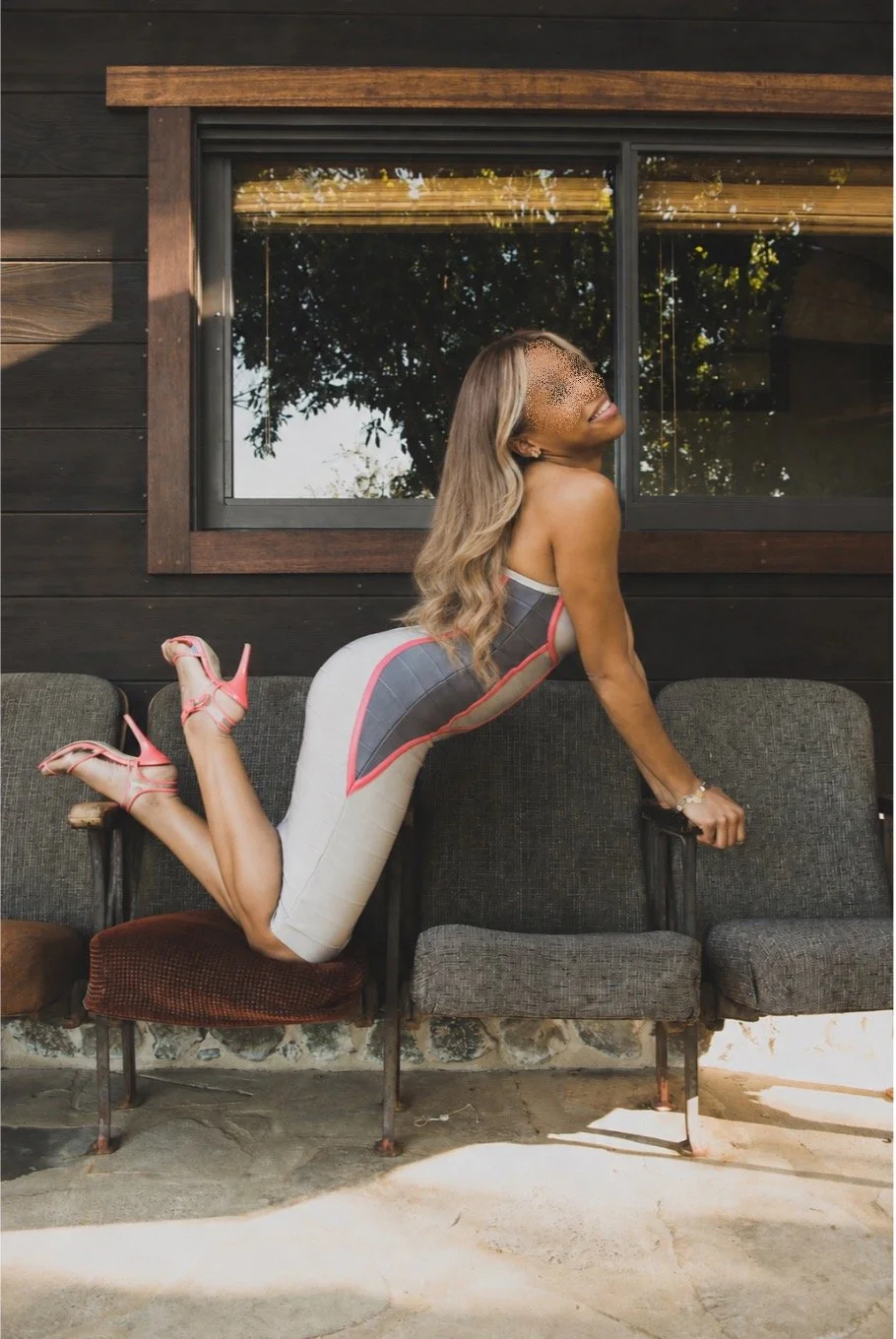 Woman in a fitted gray and pink outfit kneeling across vintage chairs outdoors.