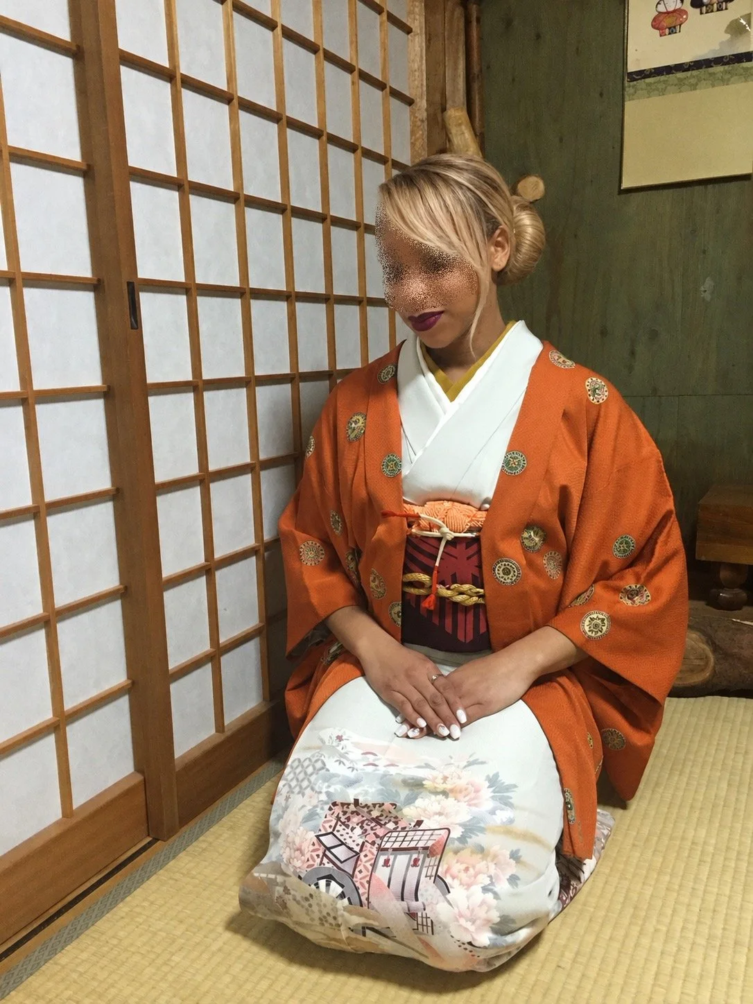 Woman wearing an orange patterned kimono seated on tatami flooring in a traditional Japanese room.