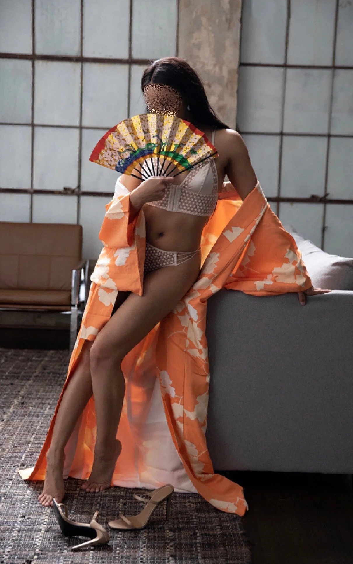 A woman in lingerie and a floral robe holding a decorative fan, standing indoors with one high heel shoe on the floor, in front of a gray sofa.