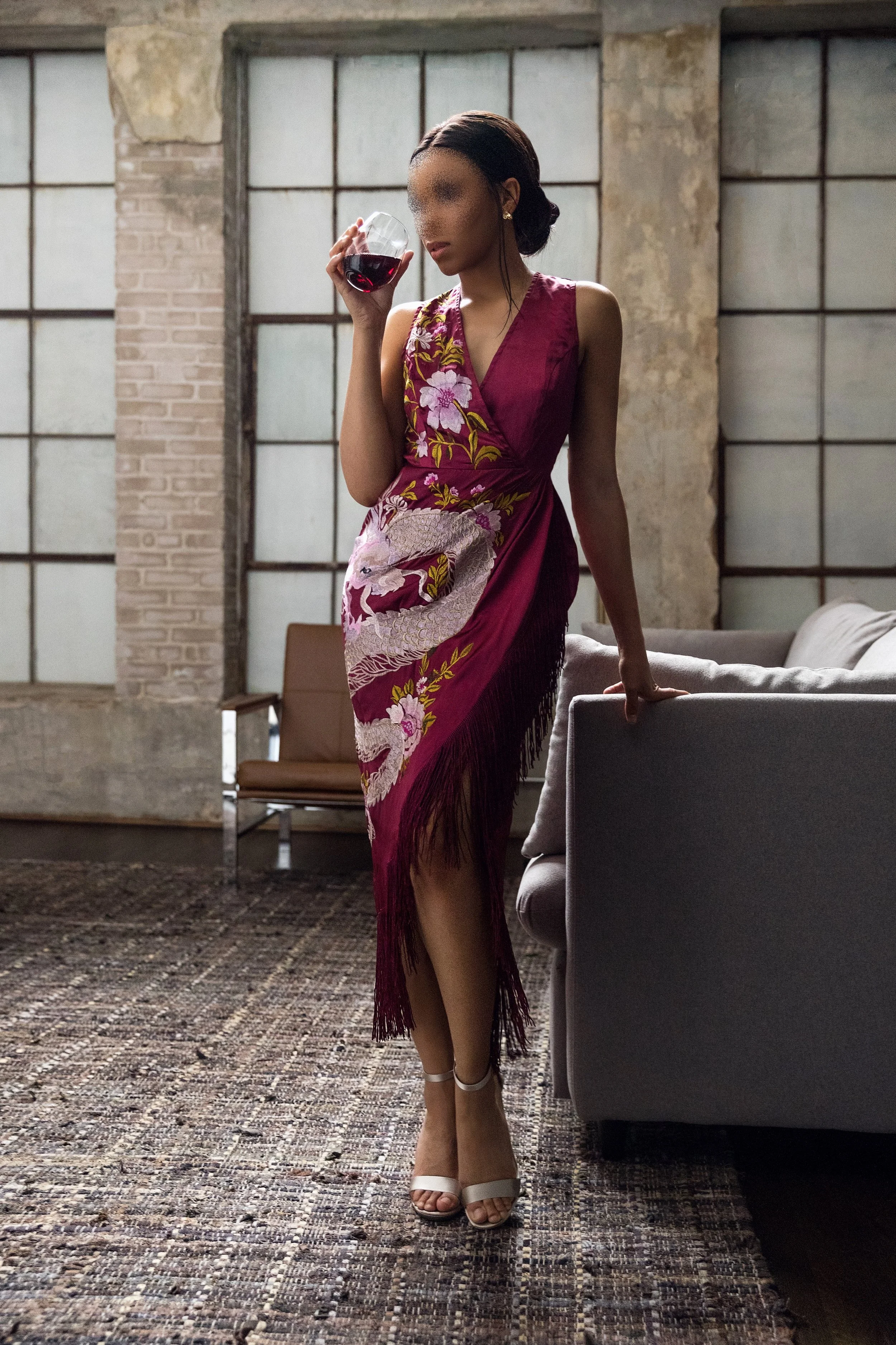 Woman in a pink floral dress and white high heels holding a glass of red wine indoors.