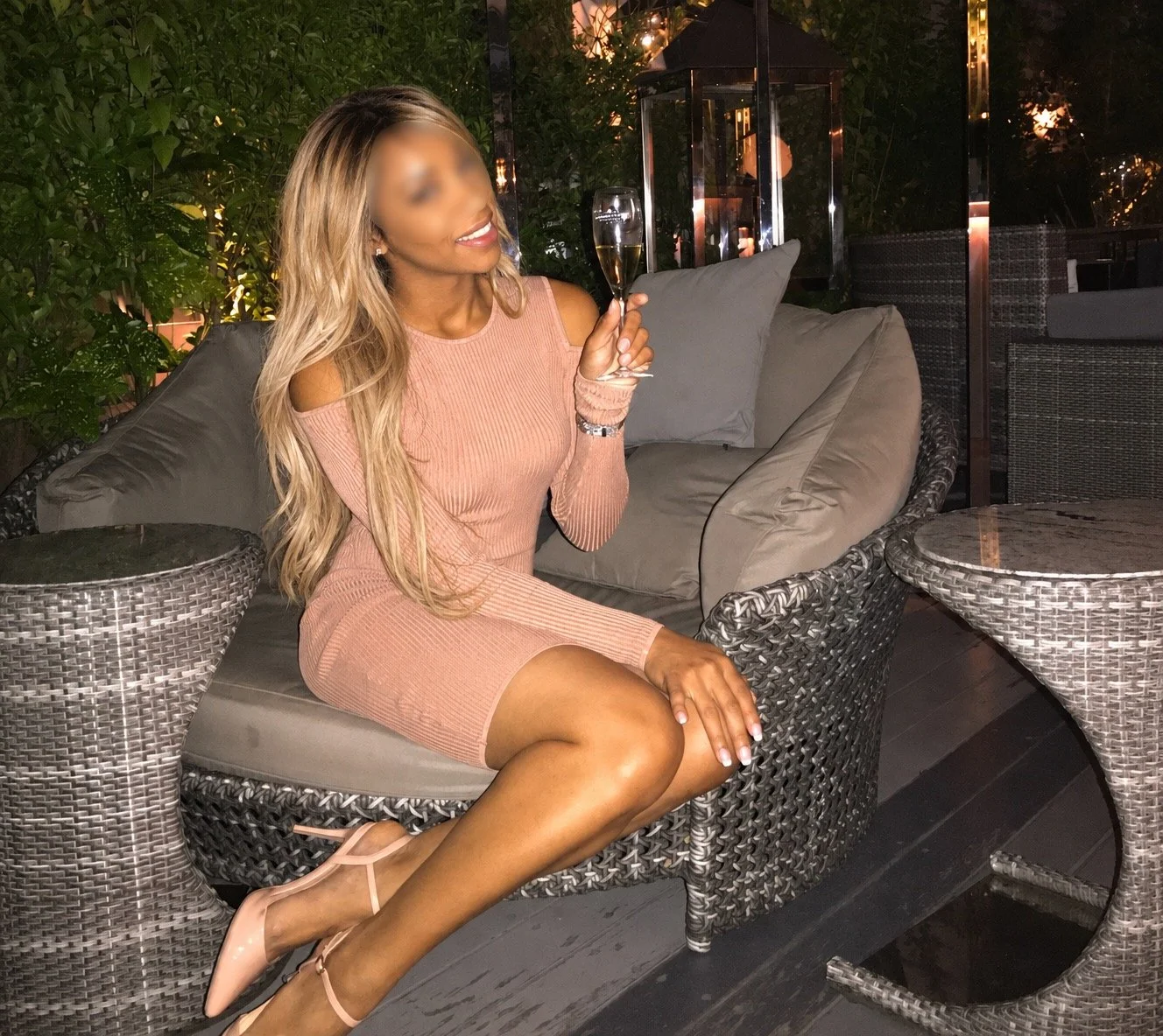 Woman in a blush pink dress holding a champagne flute while seated on an outdoor wicker lounge chair at night