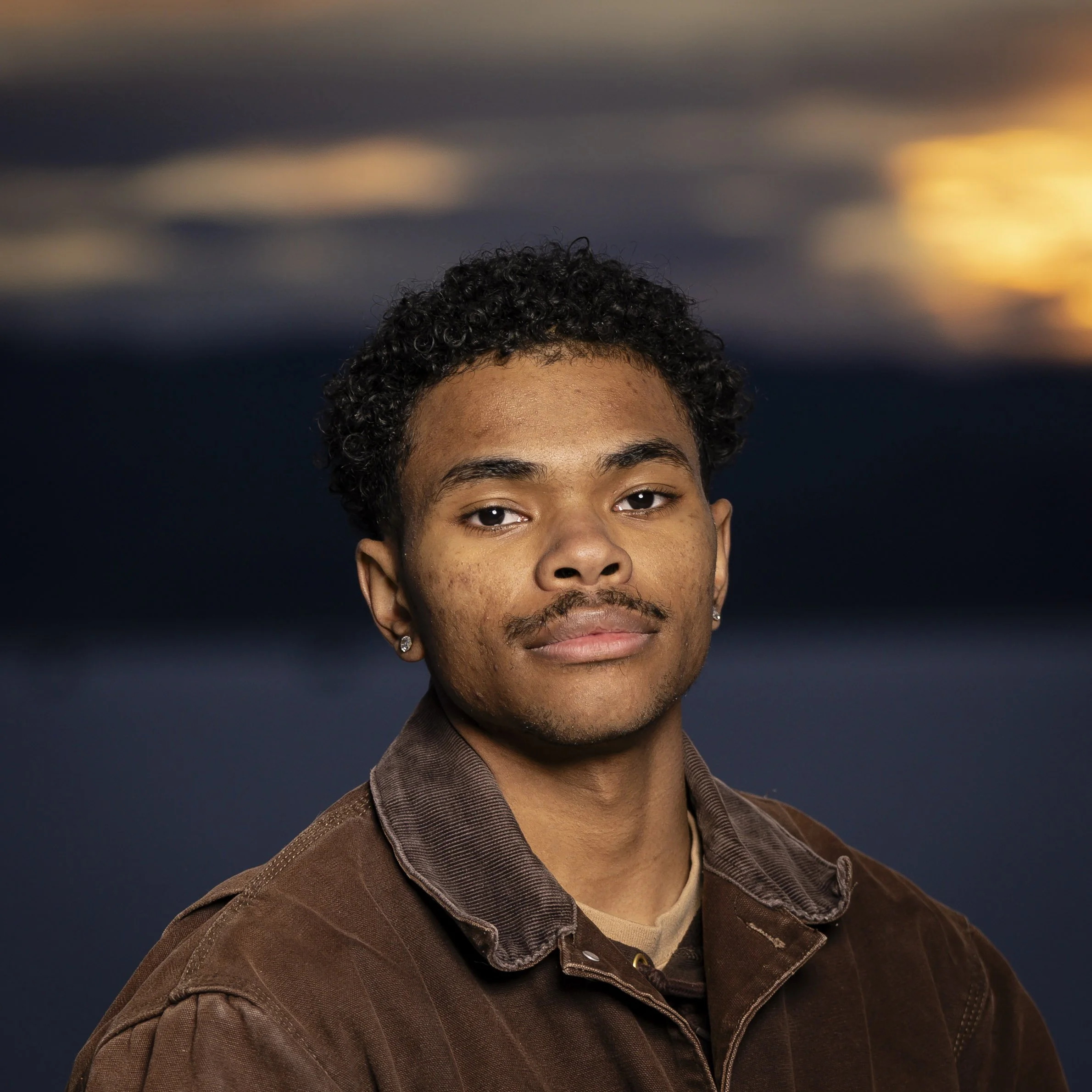 Portrait of a young man with short curly hair, mustache, earrings, wearing a brown jacket, against a dark, cloudy sky with hints of sunset in the background.