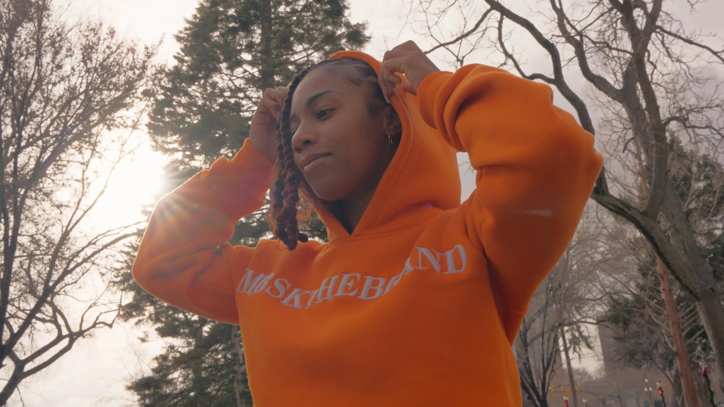 A woman adjusting her orange hoodie outdoors with leafless trees and a cloudy sky in the background.