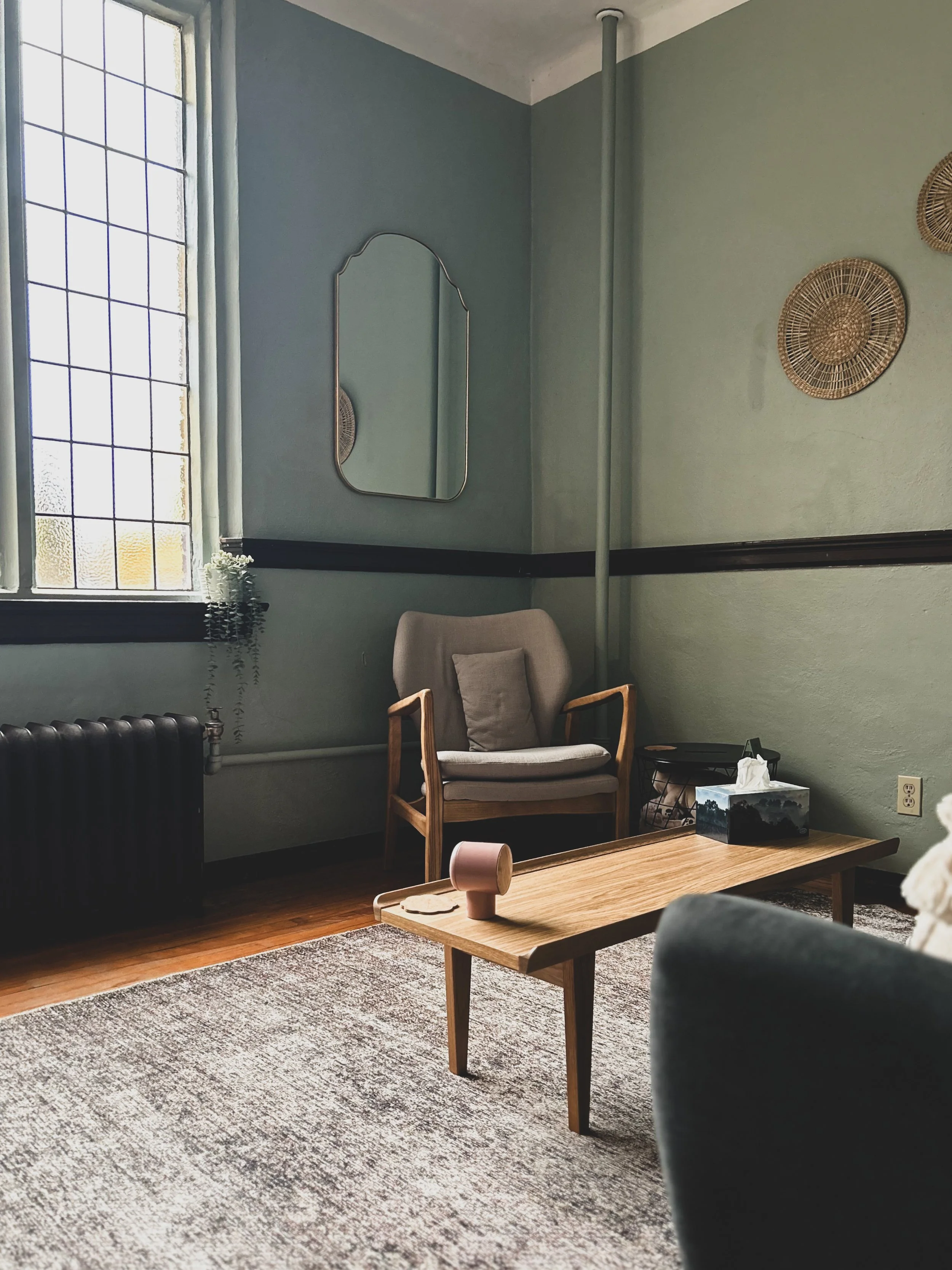 A cozy living room corner with a wooden armchair with beige cushions, a small wooden coffee table with a pinkish lamp, a tissue box, and a black side table, against pastel green and blue walls decorated with woven wall hangings and a mirror, sunlight coming through a stained glass window.