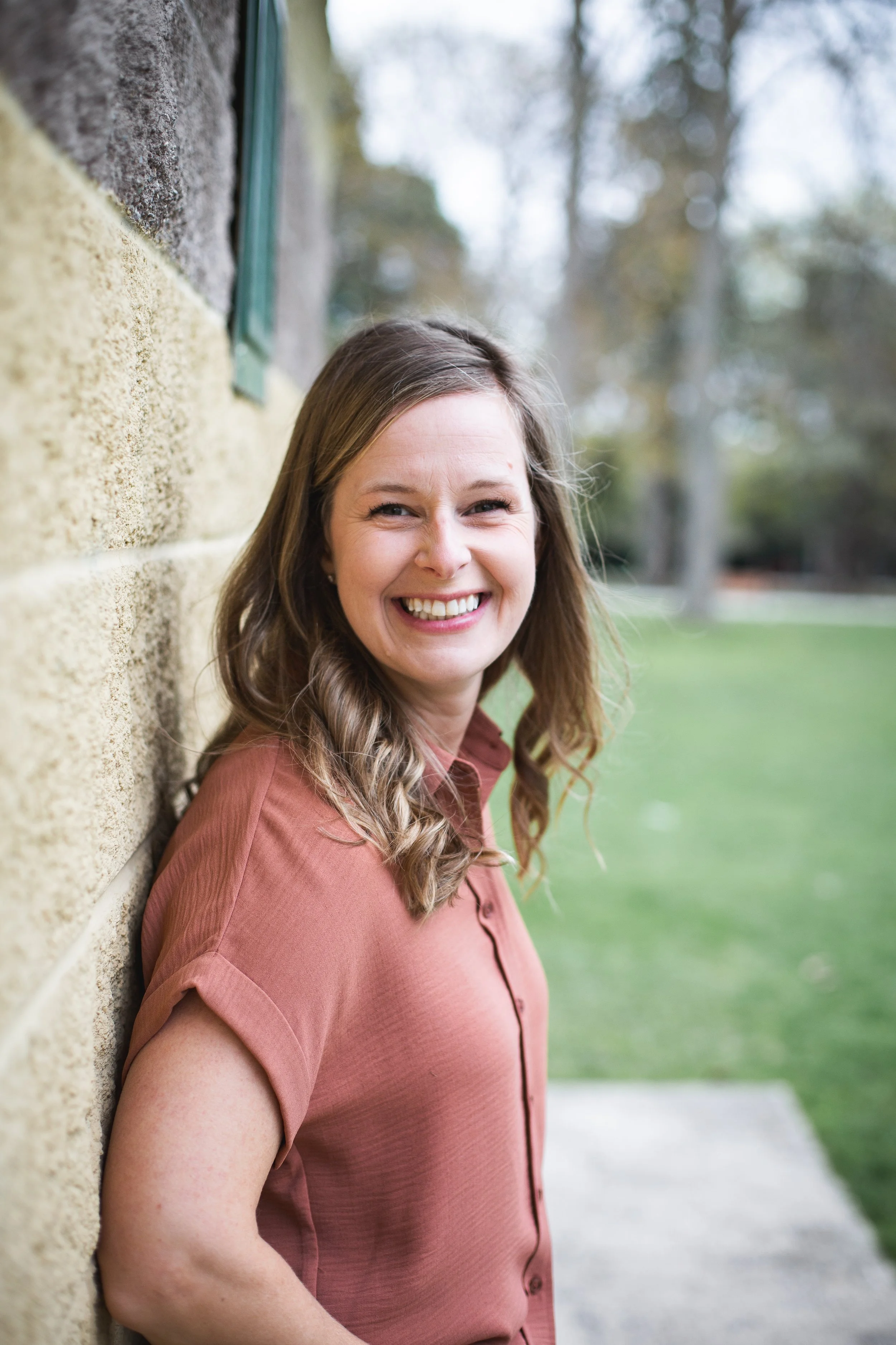 A woman with shoulder-length brown hair, wearing a rust-colored shirt, smiling and leaning against a stone wall outdoors with grass and trees in the background.