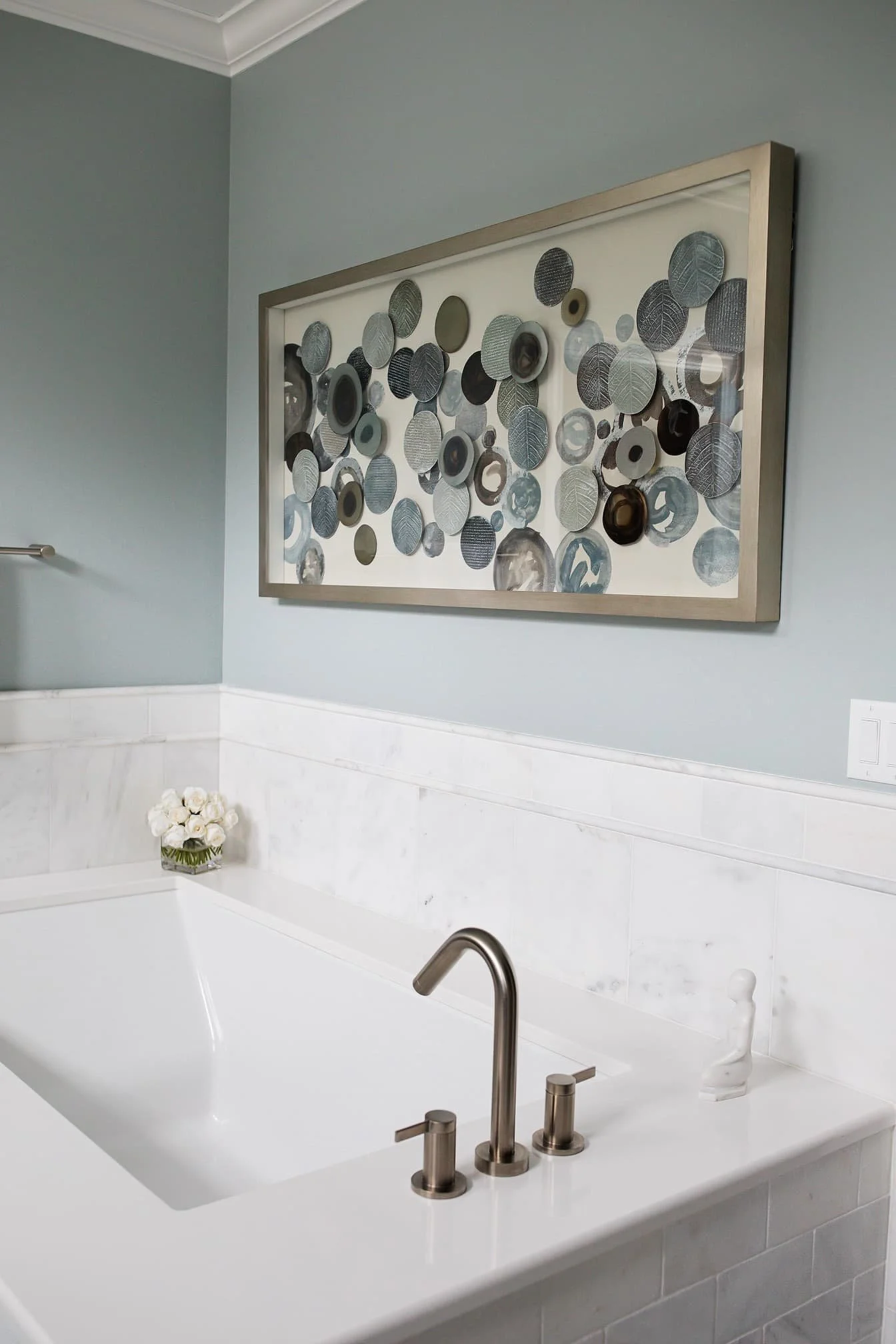Freestanding white soaking tub with brushed nickel fixtures, marble tile surround, soft blue walls, and framed modern abstract wall art in a bright, serene bathroom.