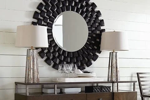 A modern sideboard with a black decorative round mirror above it, flanked by two lamps on either side, with wine glasses and books on the sideboard, in a white-walled room.