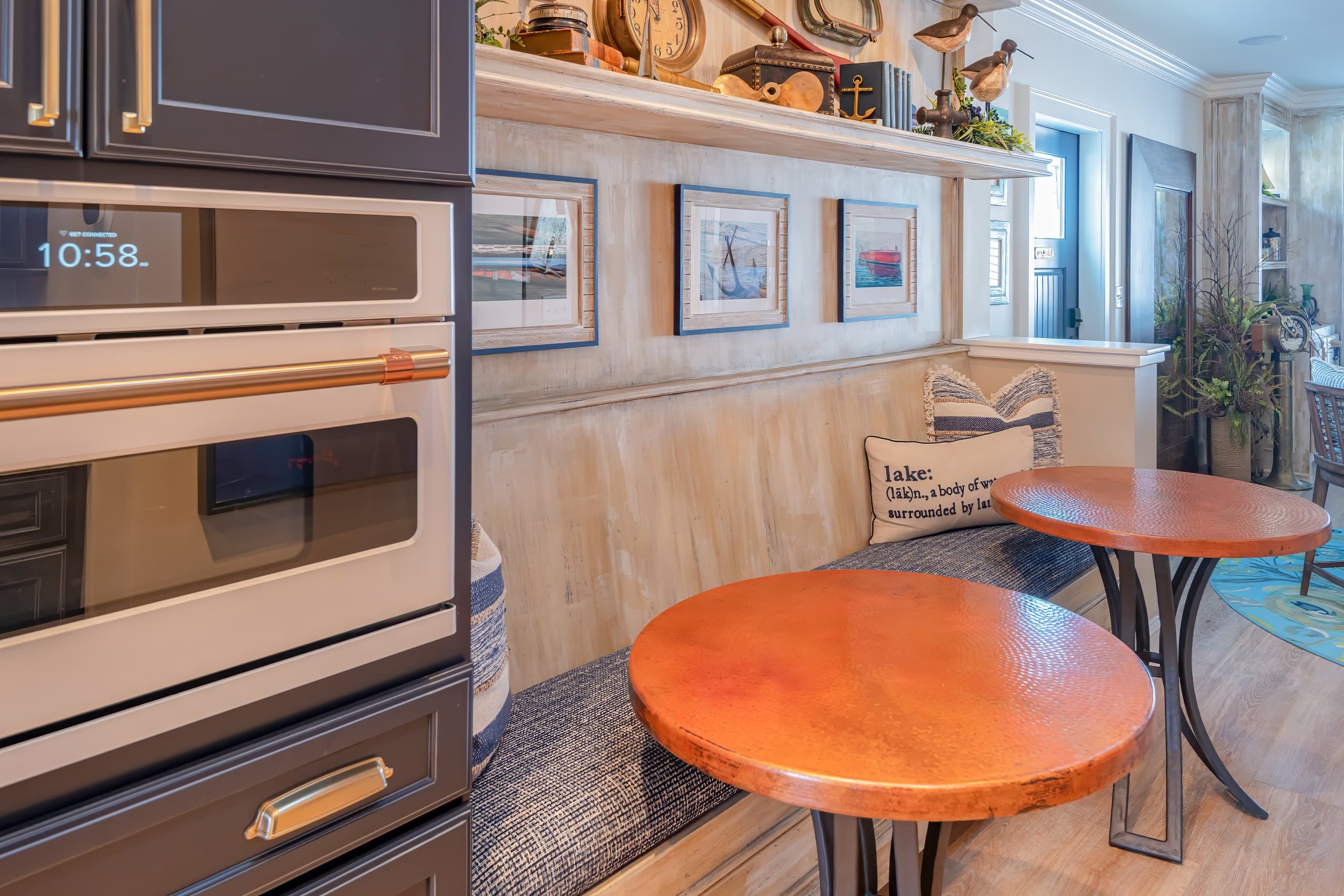 Built-in kitchen banquette with textured cushion, round hammered metal tables, coastal artwork, and nautical decor displayed on a shelf above.