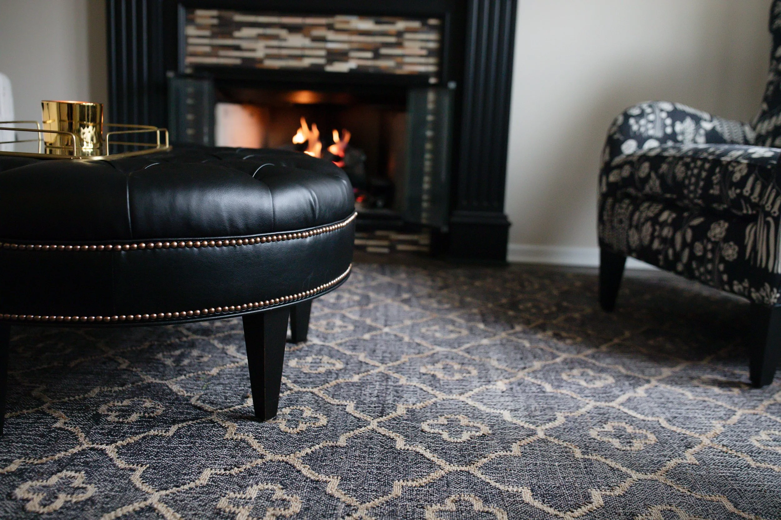Detail view of custom leather ottoman, patterned area rug, and fireplace, highlighting layered textures and refined finishes in a sophisticated interior design.