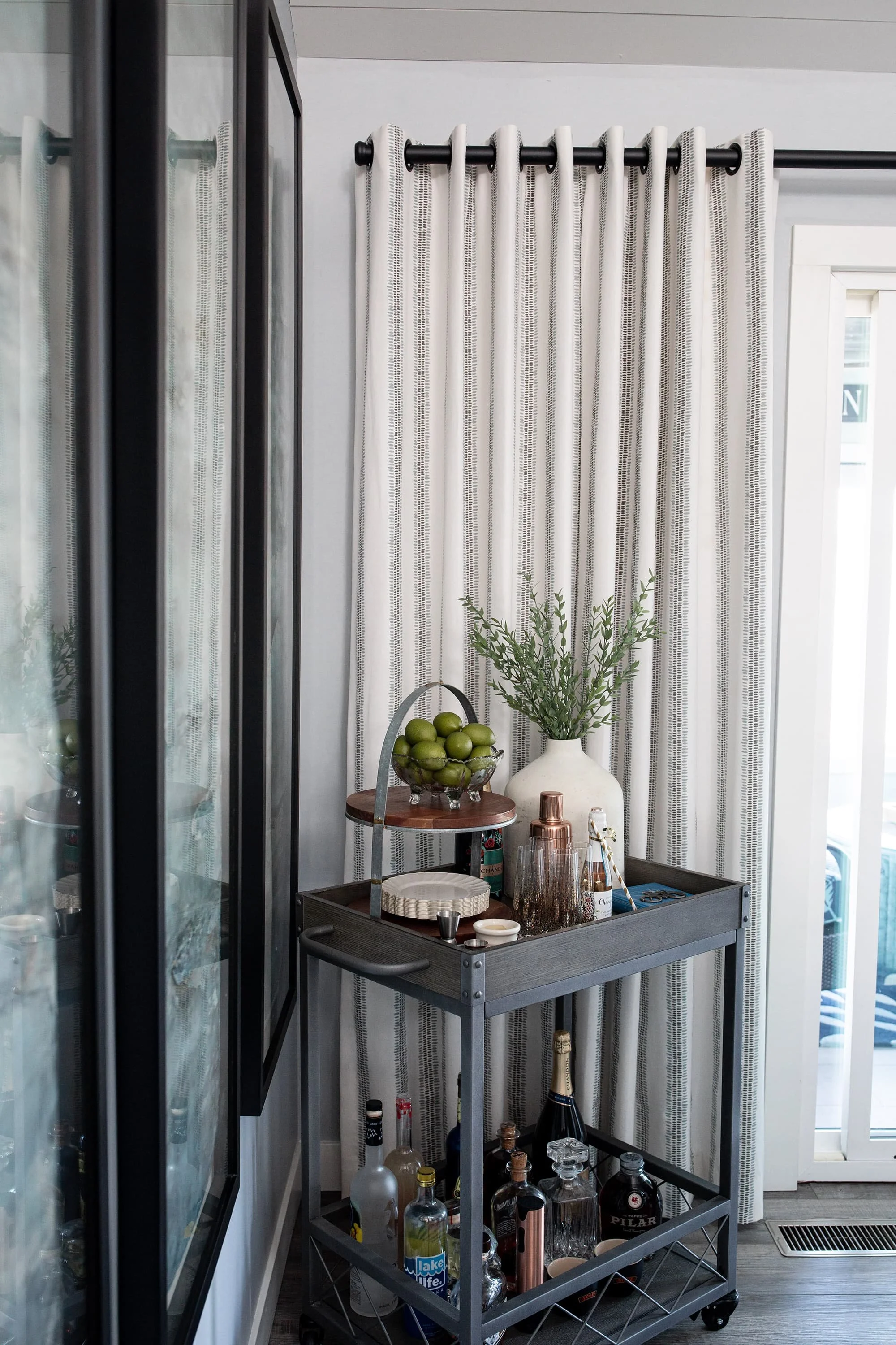 Styled bar cart with glassware, bottles, greenery, and decorative accents beside neutral drapery and a glass door in a bright, modern space.