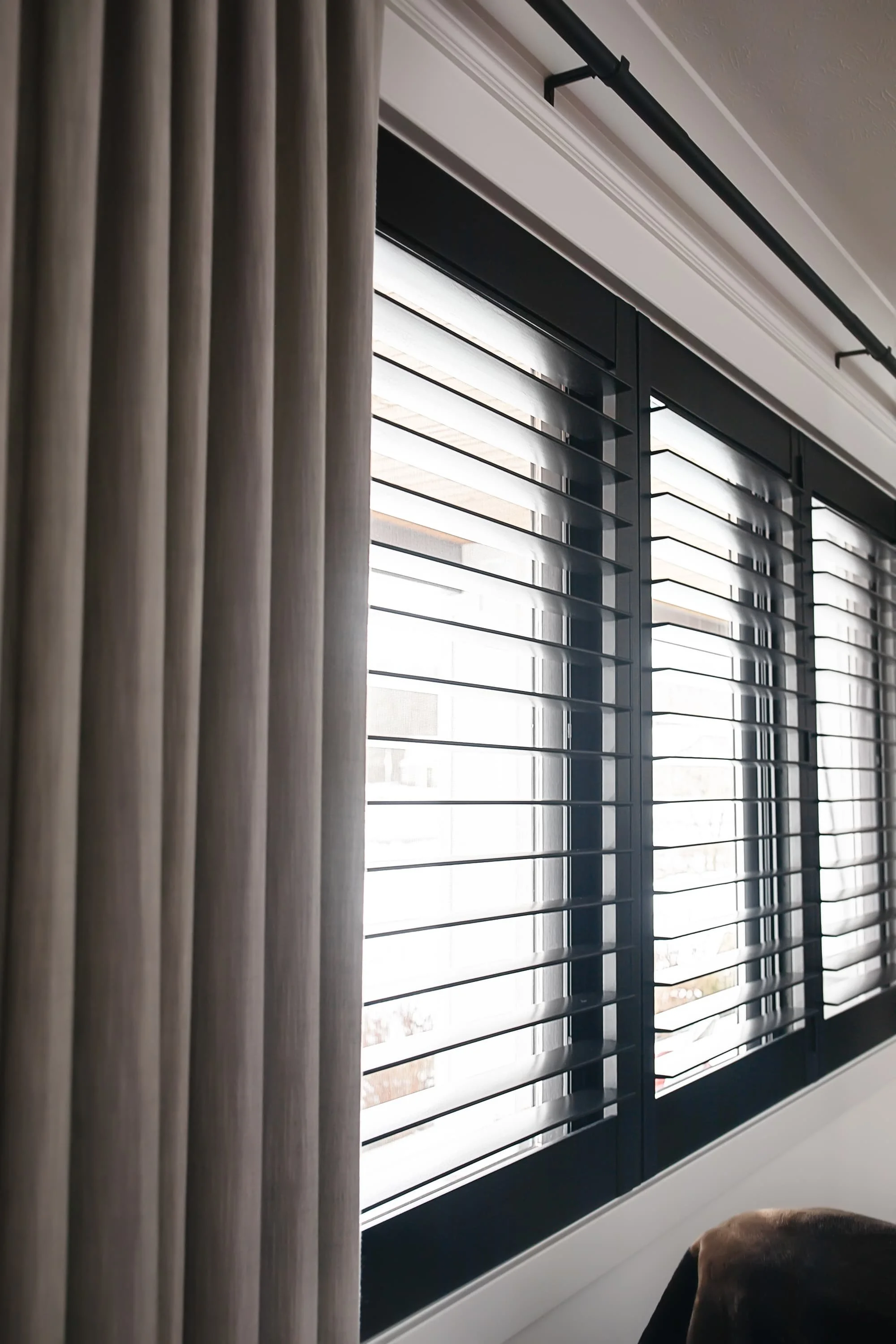 Black interior window shutters with horizontal slats paired with neutral linen drapery on a black rod, allowing soft filtered light into a modern space.