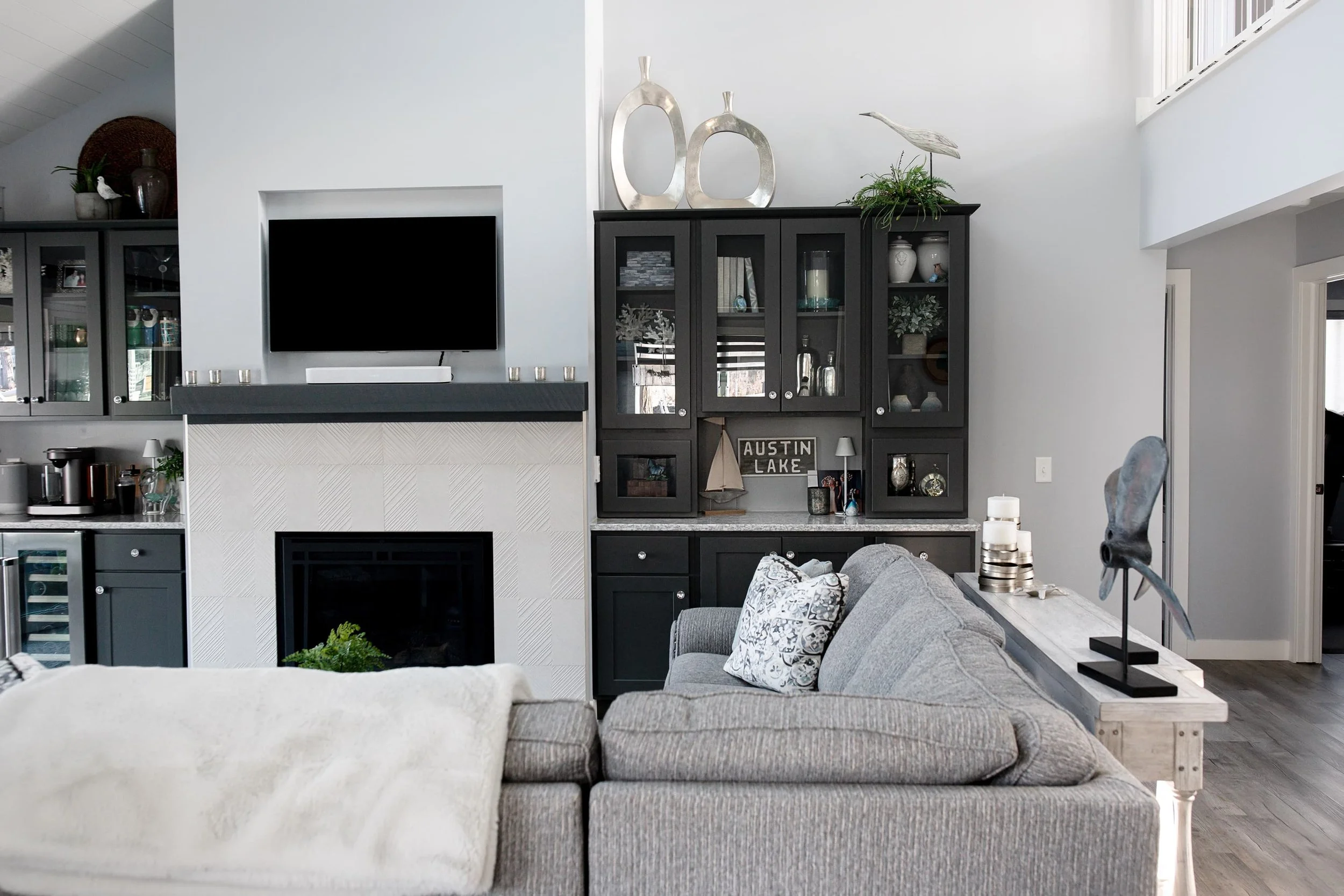 Living room with gray sectional sofa, dark built-in cabinetry, fireplace with light tile surround, mounted TV, and styled shelves with coastal-inspired decor.