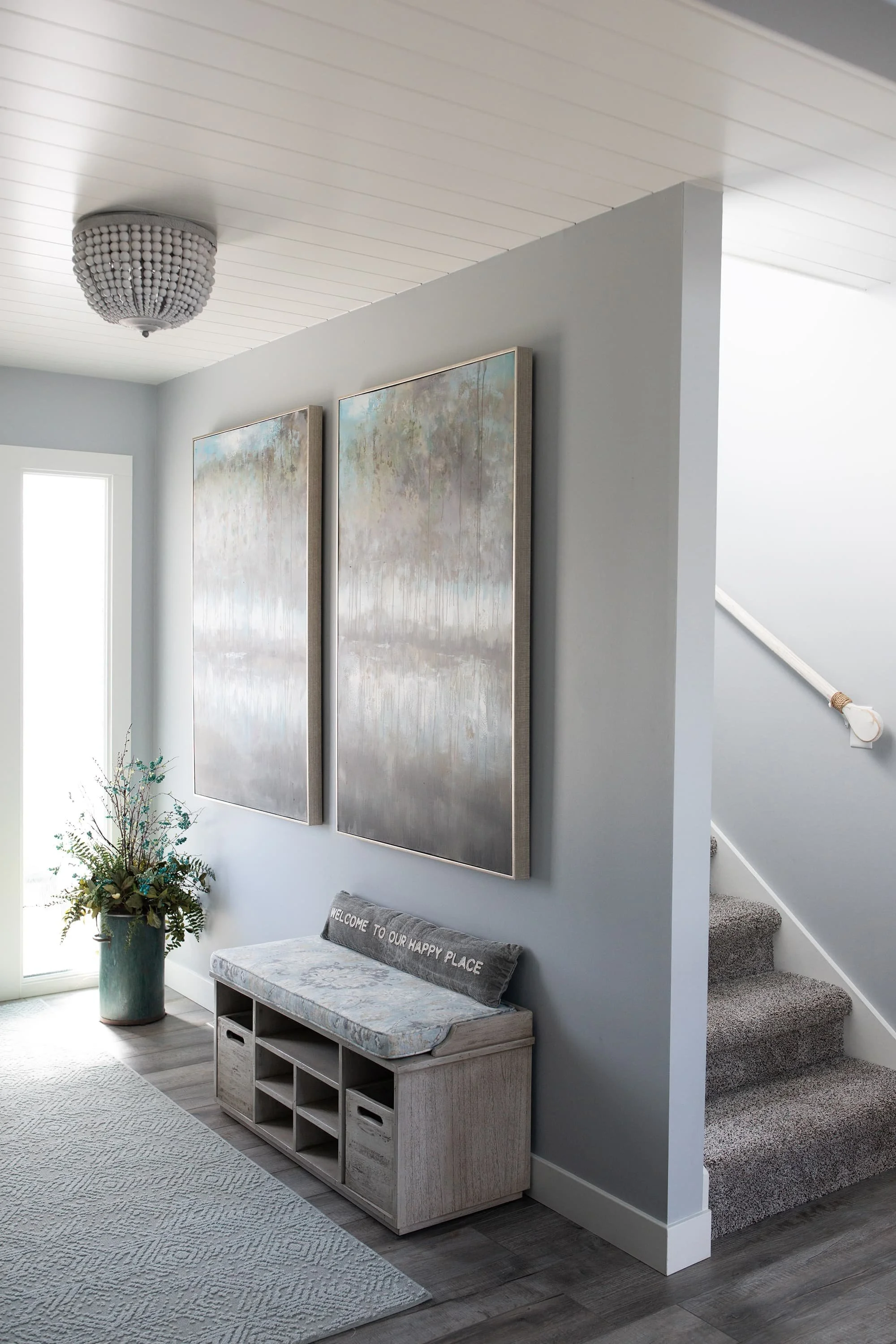 Entryway with blue-gray walls, large abstract artwork, wood bench with storage, textured rug, and soft natural decor creating a calm, welcoming space.