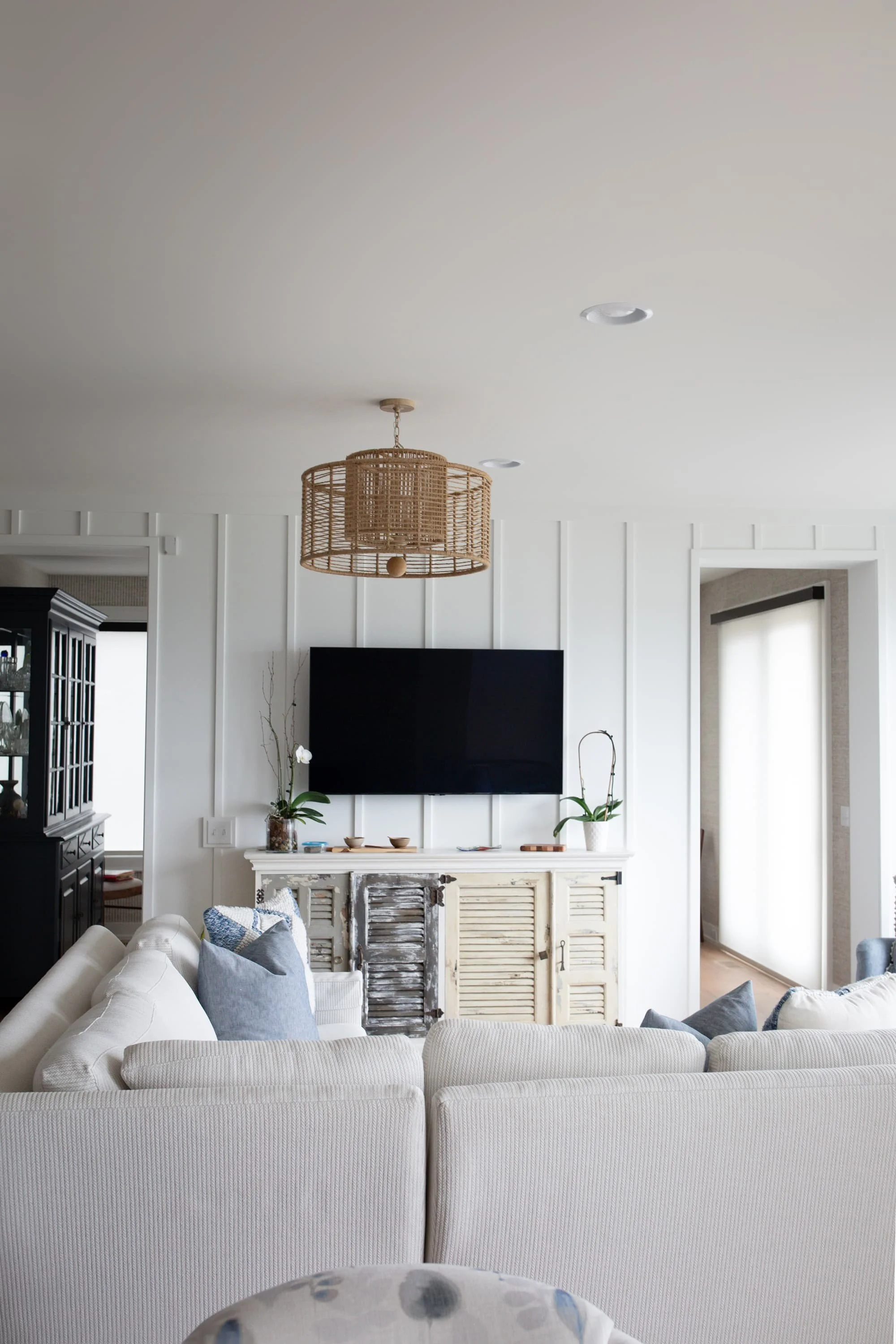 Bright living room with white paneled walls, woven pendant light, neutral sectional sofa with blue pillows, wood console, and soft window shades.