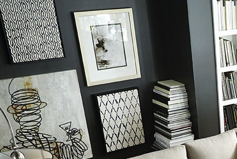 A wall with various monochrome framed art pieces and a shelf filled with stacked books, part of a living room or office decor.