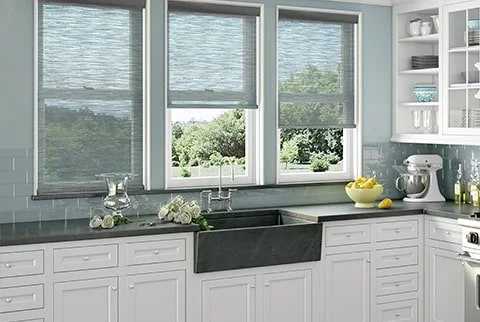 Kitchen with large windows, white cabinets, gray countertop, and a farmhouse sink. Items on the counter include a glass pitcher, white flowers, a yellow bowl with lemons, and a stand mixer.