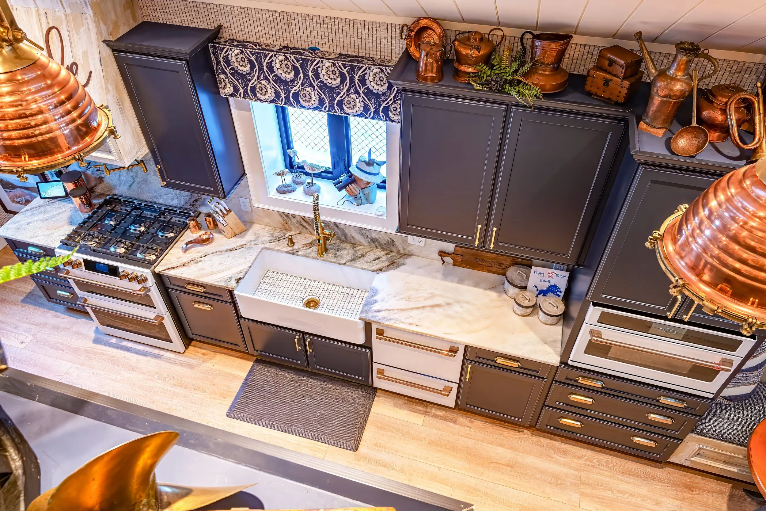 Overhead view of a kitchen with navy cabinetry, marble countertops, a farmhouse sink, brass fixtures, and copper range hoods, with warm wood flooring and layered nautical-inspired decor.