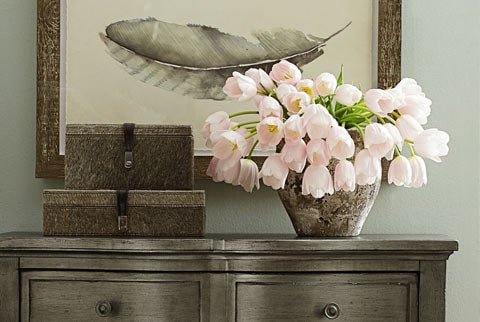 Decorative arrangement with pink flowers in a vase, a framed leaf print on the wall, and three small storage boxes on a dark wooden dresser.