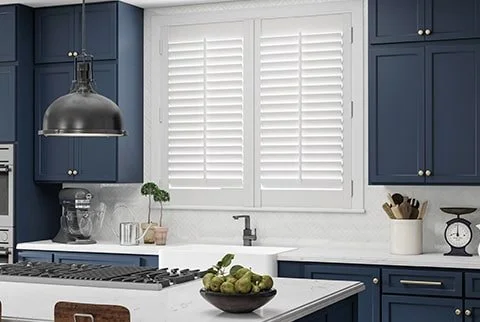 Modern kitchen with blue cabinets, white subway tile backsplash, white countertop, window with white shutters, and kitchen appliances.