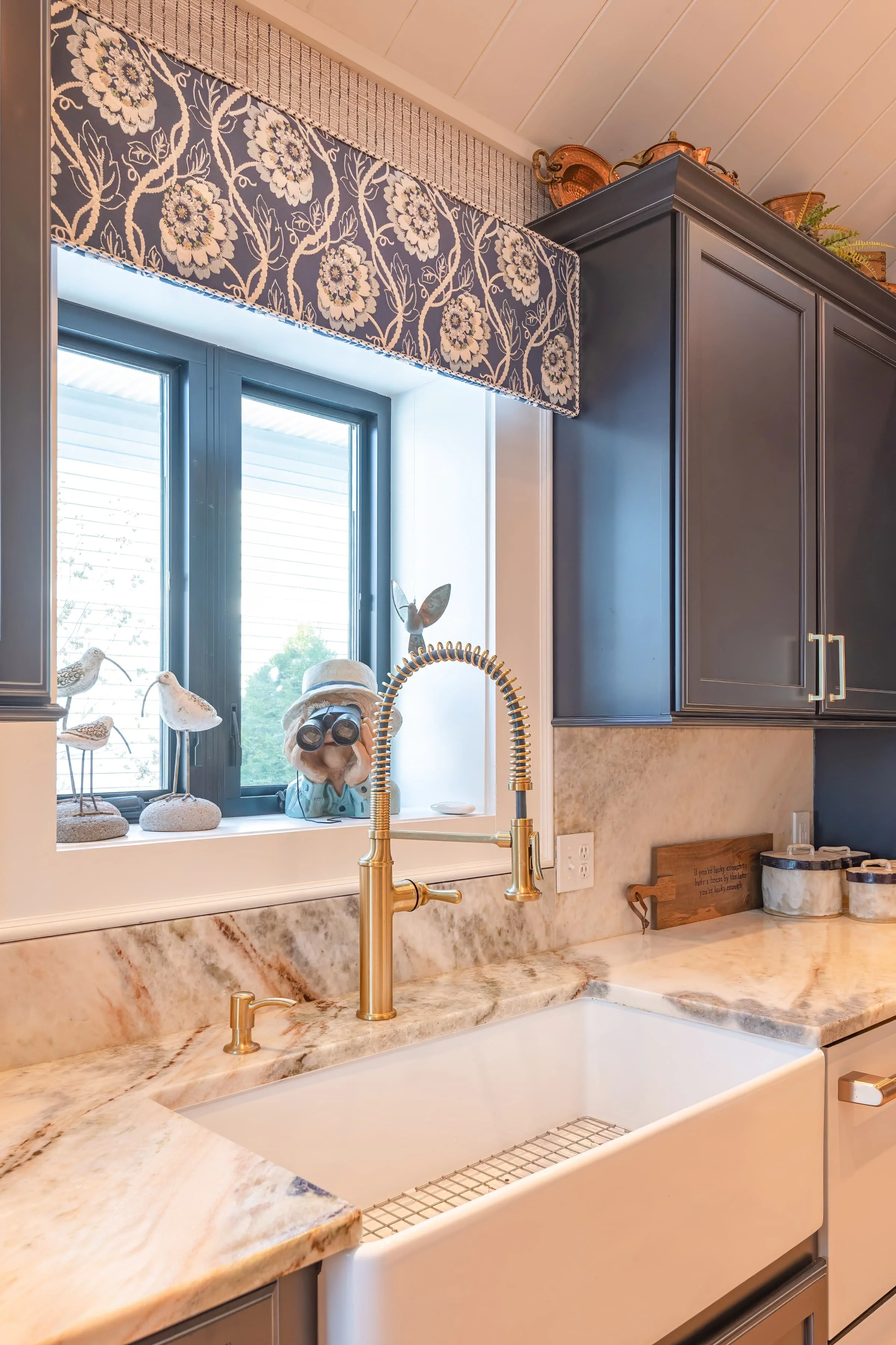 Kitchen with navy cabinets, brass gooseneck faucet, farmhouse sink, marble countertops, and patterned blue valance above a window with coastal decor accents.