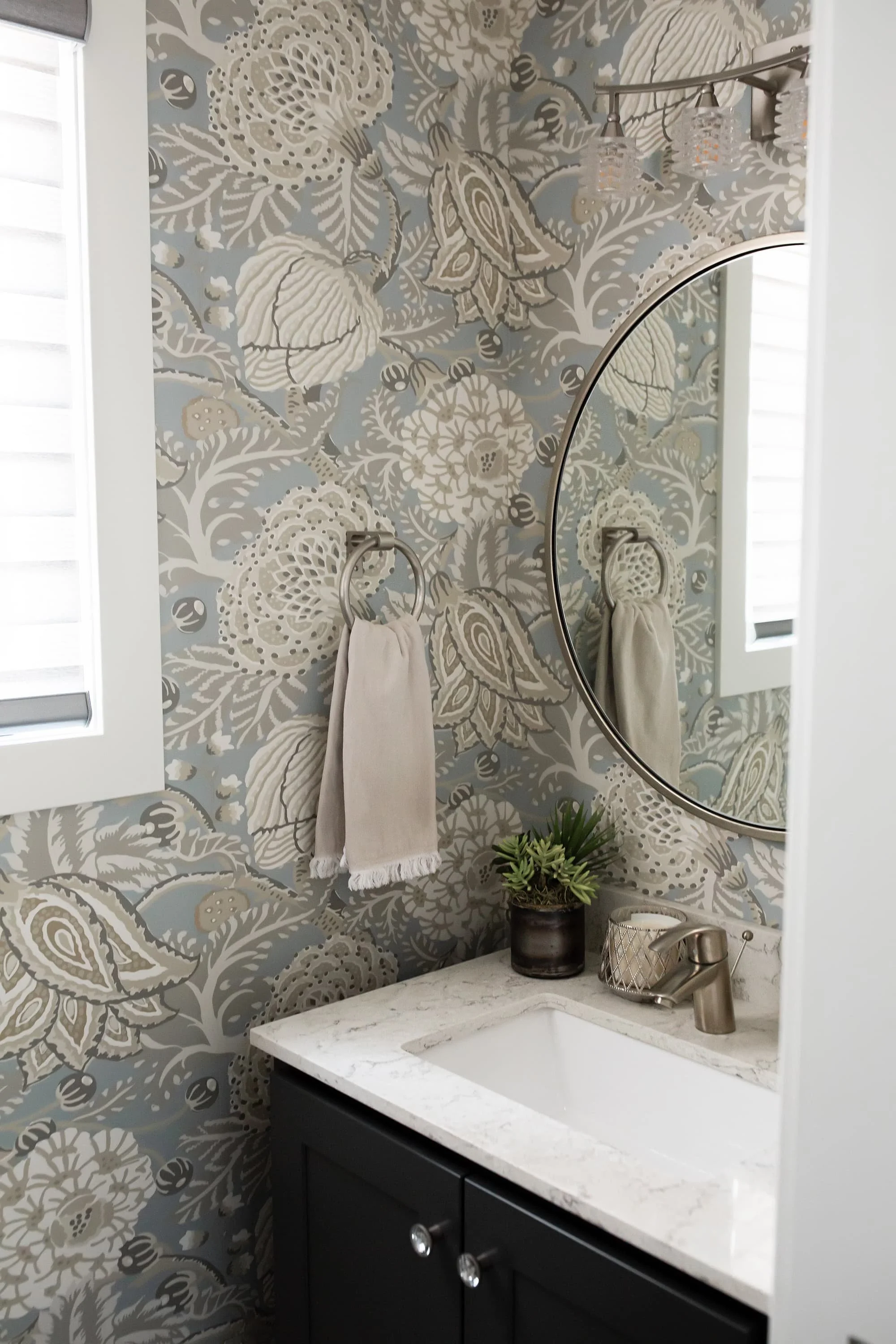 Powder bathroom with blue and neutral floral wallpaper, marble countertop, dark vanity, round mirror, and brushed metal fixtures in a refined, modern space.